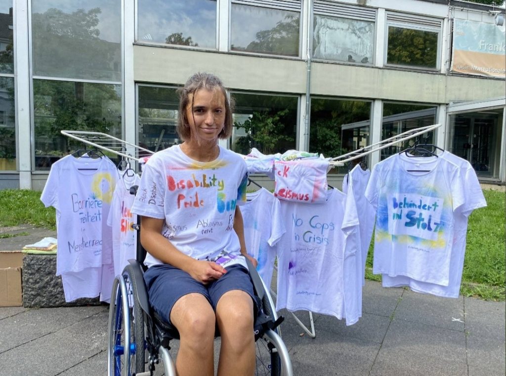 Foto von Lena, einer Orga Person der Disability und Mad Pride Bonn, vor einem Wäscheständer mit selbst gestalteten weißen T-shirts.