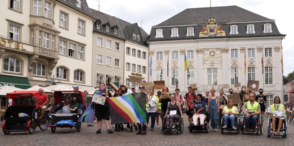 Gruppenfoto der Teilnehmer*innen an der Laufdemo der Disability und Mad Pride Bonn vor dem Bonner Rathaus. Zu sehen sind die Teilnehmer*innen, eine große Disability Pride Flagge, Demoschilder und Rikschas, außerdem das Rathaus.