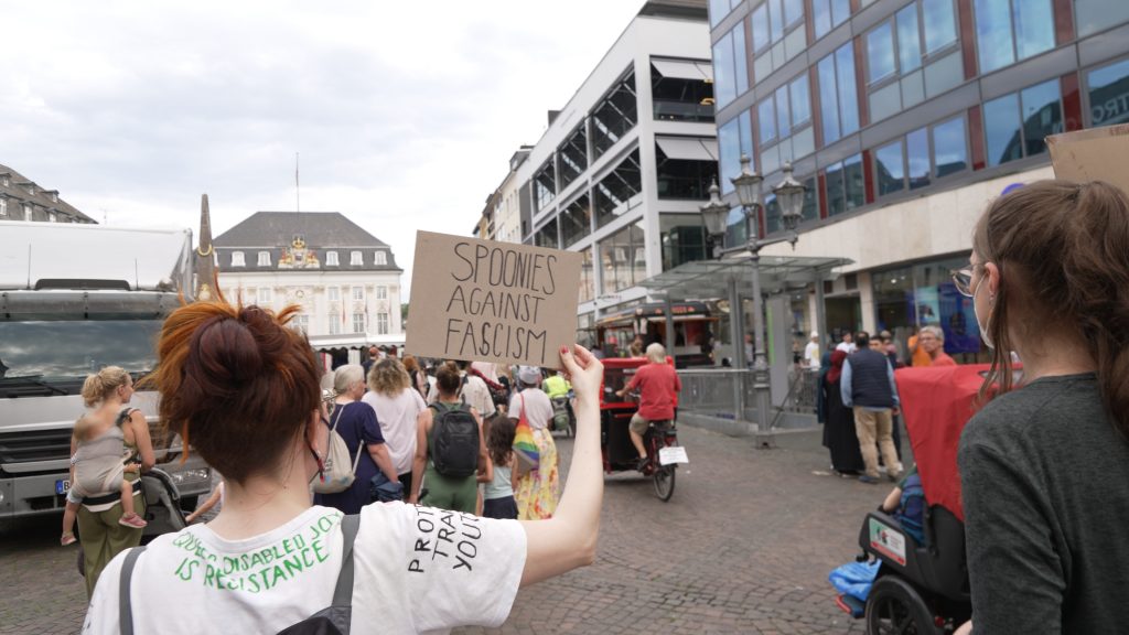 Foto einer Person von hinten mit weißen T-shirt und einem Demoschild. Auf dem T-shirt sind die Schriftzüge "Queer disabled joy is resistance" und "Protect trans youth" zu lesen. Auf dem Schild steht "Spoonies against fascism".